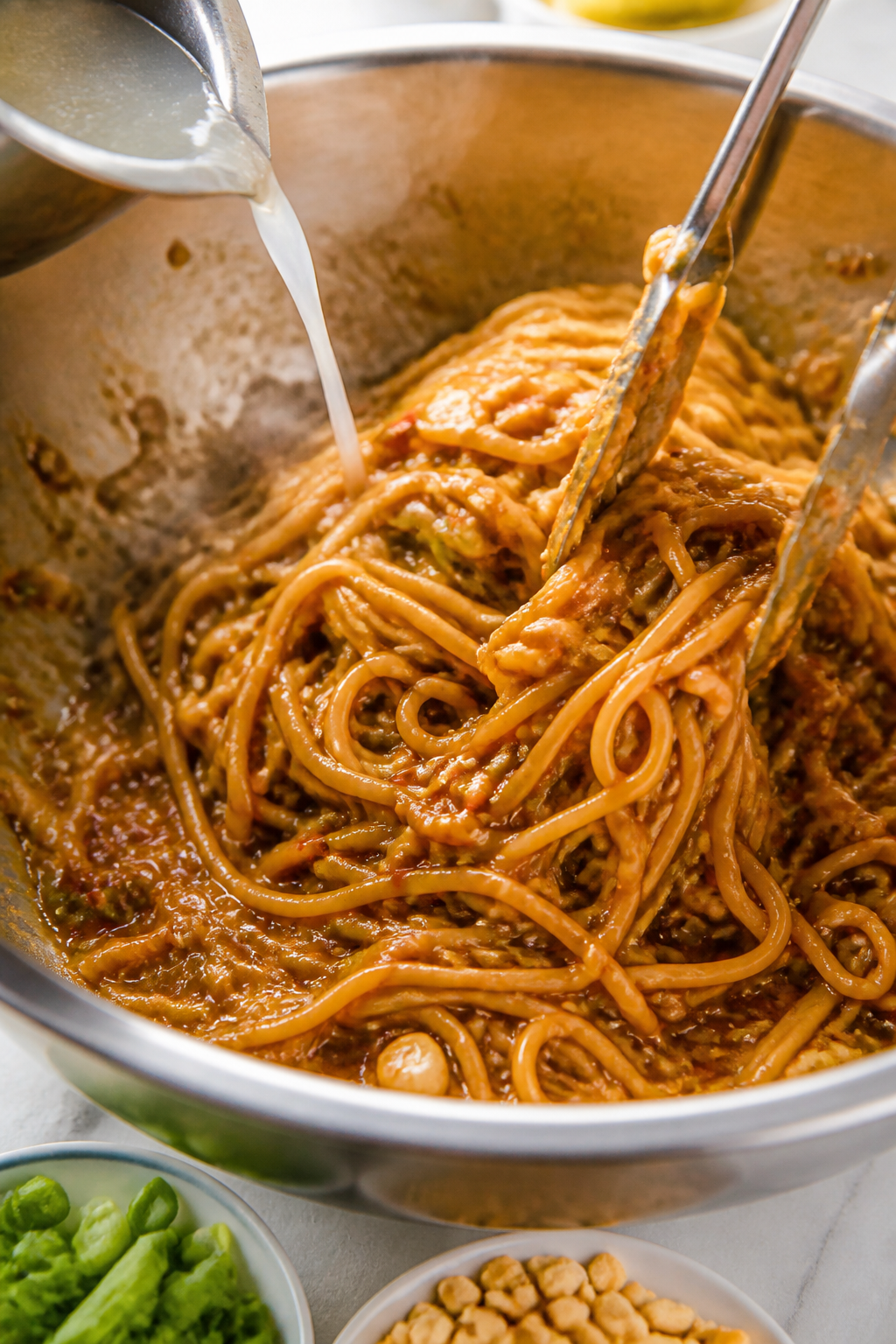 Cooking process close-up: Hot lo mein noodles being tossed in a large stainless bowl with a glossy p